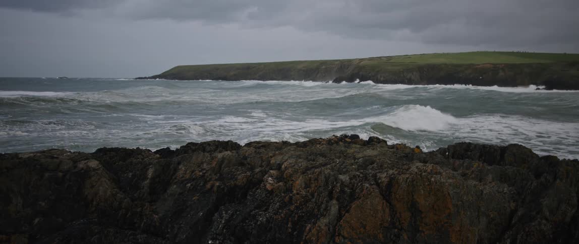 A static wide shot of a rugged, rocky coastline as waves from a choppy sea roll towards the shore under a grey, overcast sky