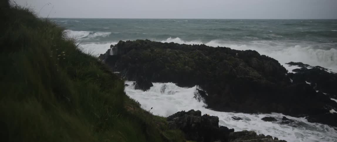 Dramatic slow motion shot of a stormy ocean with large waves crashing over mossy rocks from a grassy cliff