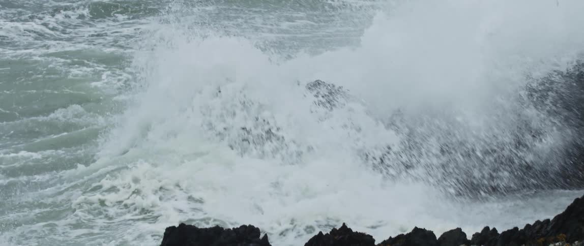 A powerful ocean wave crashes against dark jagged rocks in dramatic slow motion