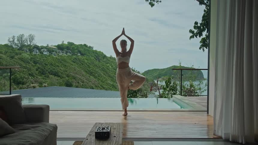 A woman holds a tree yoga pose on a wooden deck next to an infinity pool overlooking a tropical hillside and the ocean