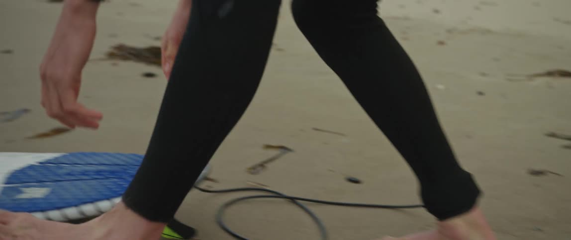 Close up of a surfer attaching their leash and picking up their surfboard on a sandy beach
