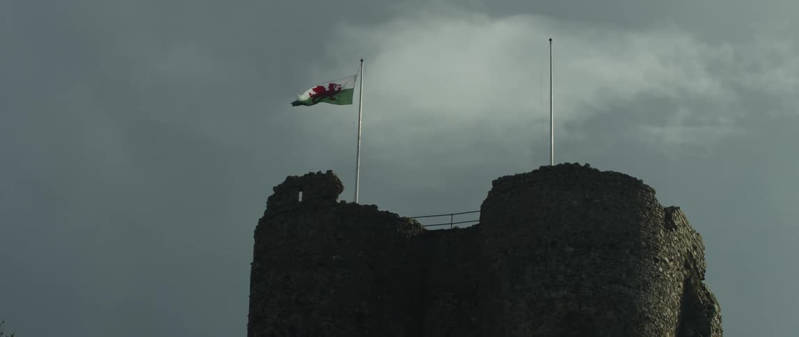 The Welsh flag flies at half-mast in the wind atop a stone castle ruin under a dramatic cloudy sky