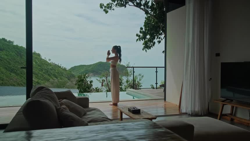 A serene woman practices yoga and meditates on the deck of a luxurious tropical villa overlooking a beautiful ocean and hillside view