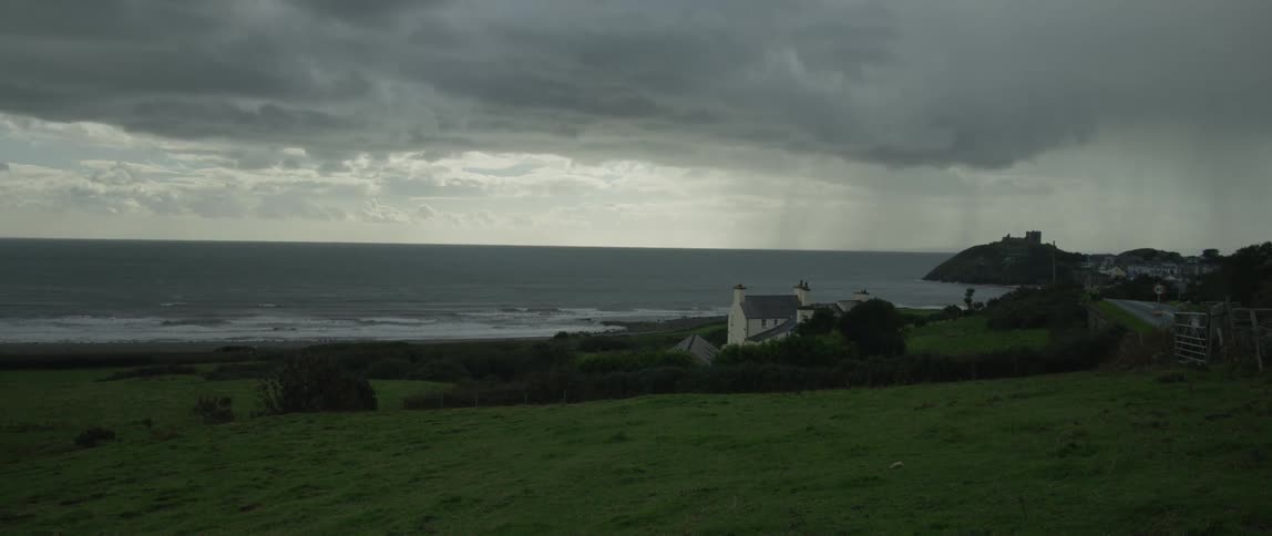 Rain falls over the ocean on a moody day, viewed from a green hill overlooking a coastal home and a distant castle