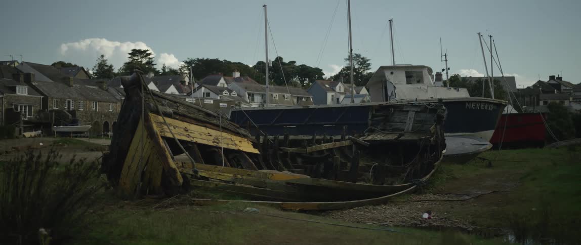 The skeletal remains of an old wooden boat lie abandoned on the grassy shore of a coastal harbor village