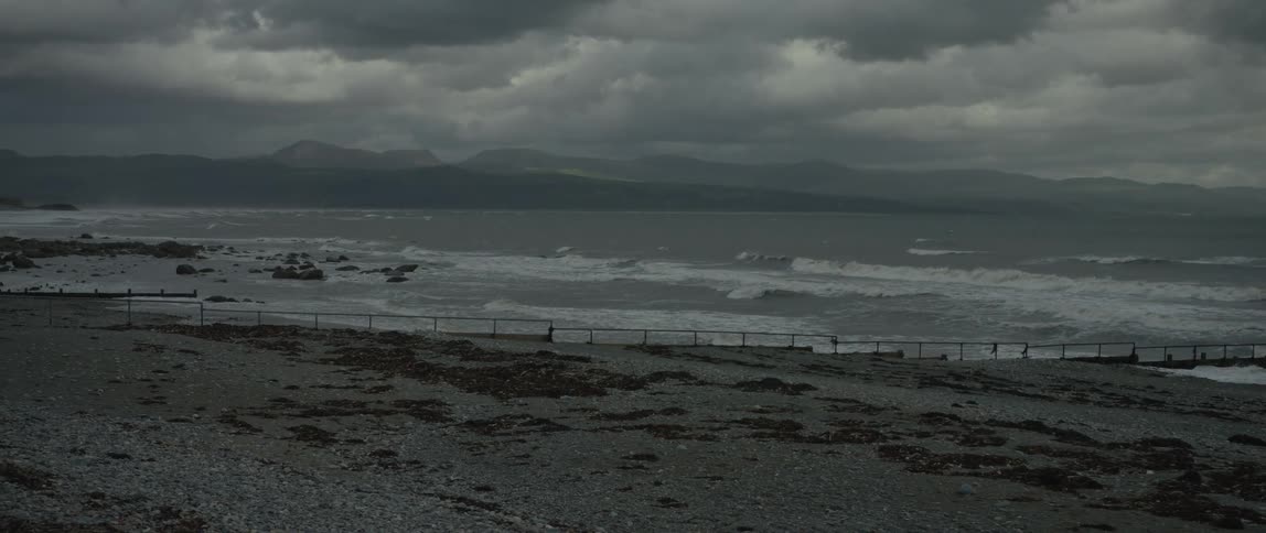 A wide, static shot shows a turbulent ocean with white-capped waves crashing against a rocky shore under dark, stormy clouds, with mountains in the distance