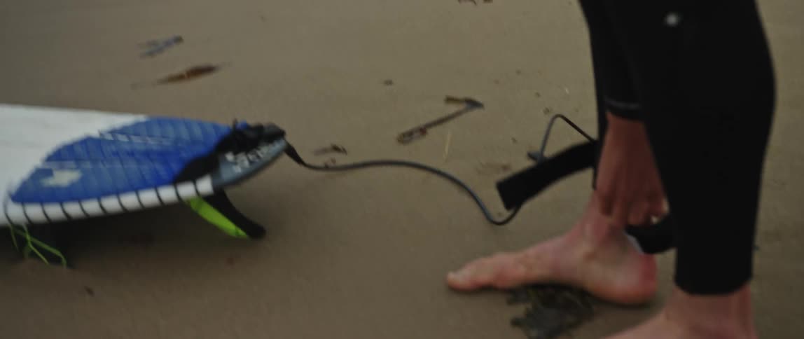 Close-up of a surfer in a wetsuit strapping a leash to their ankle on the sand before heading out to sea