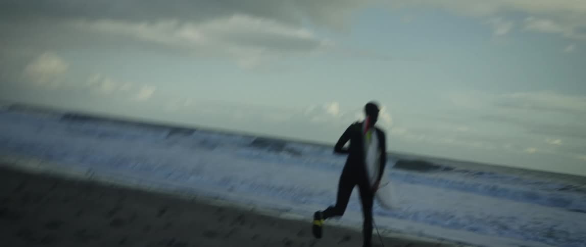 A determined surfer in a full wetsuit runs with a surfboard in slow motion along the beach and into the ocean waves at dusk