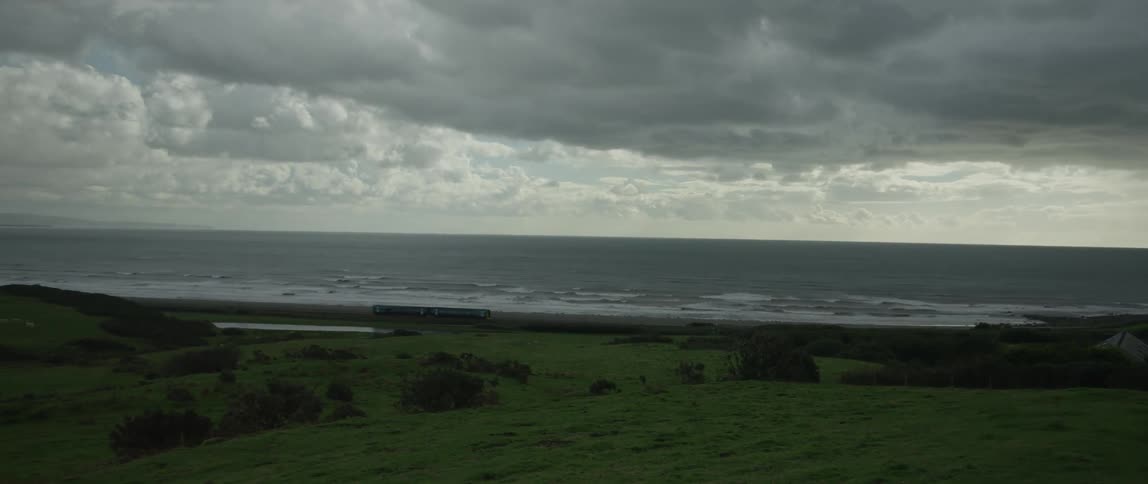 A scenic view of a passenger train traveling along the green, moody coastline of the United Kingdom under a dramatic cloudy sky
