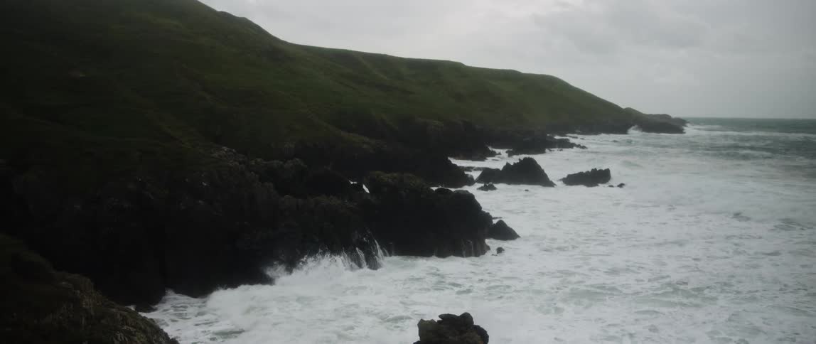 Dramatic slow motion shot of powerful white waves crashing against the dark, rocky cliffs of a green coastline