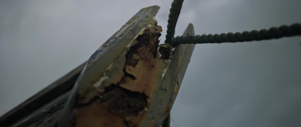 Close up of a weathered and rotting wooden boat with peeling paint tied with a rope under a gloomy sky