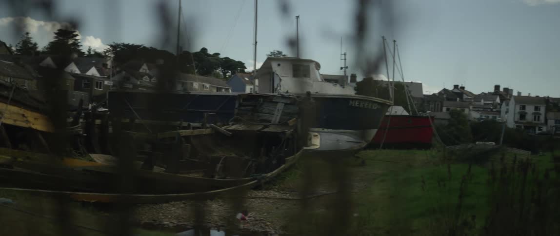 A slow pan reveals an old derelict wooden boat rotting away in a harbor amongst newer vessels, with a quaint coastal village in the background