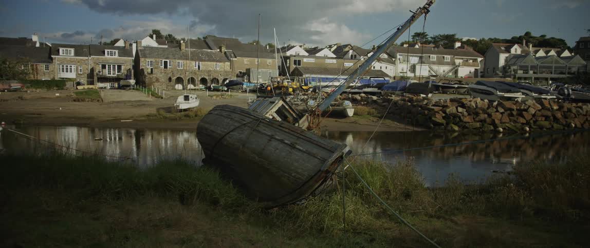 An old overturned wooden boat rests on a grassy bank in a quiet coastal harbor town