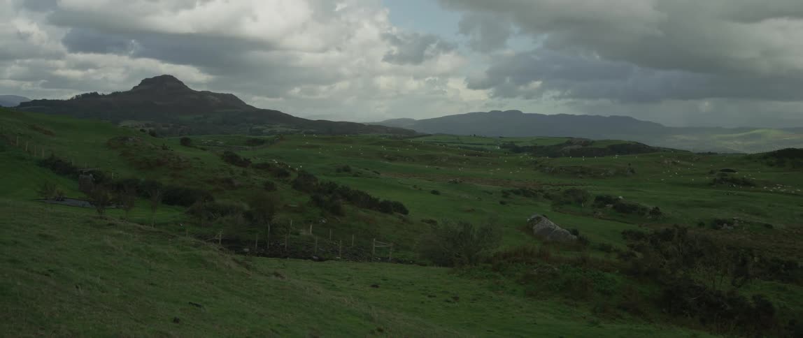 A static wide shot of a vast, rolling green countryside with distant mountains under a dramatic, cloudy sky