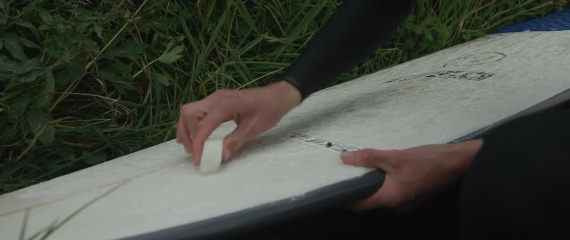 Close up of a surfer in a wetsuit applying wax to their surfboard while sitting in the grass