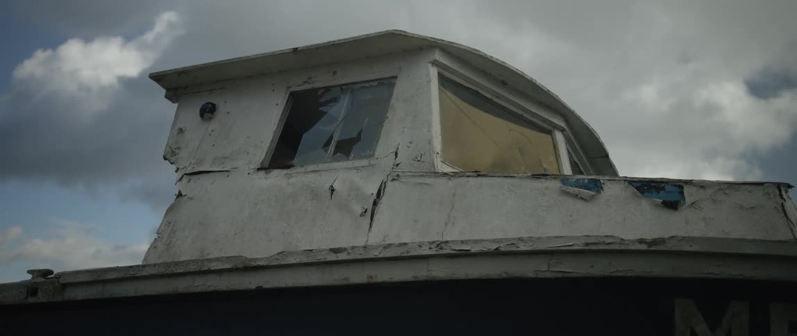 A close-up of a derelict boat's cabin with shattered windows against a moody, overcast sky