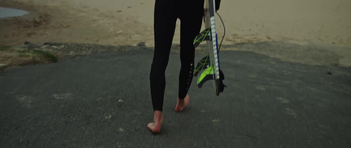 A surfer in a wetsuit walks barefoot towards the choppy ocean with his surfboard on a cloudy day