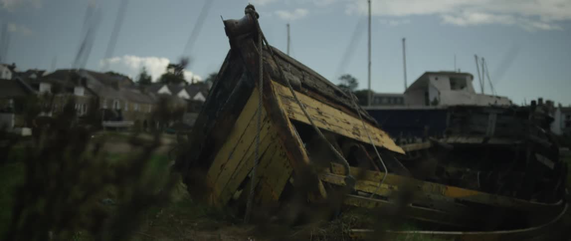 A weathered and decaying yellow wooden boat sits abandoned on the grassy shore of a harbor