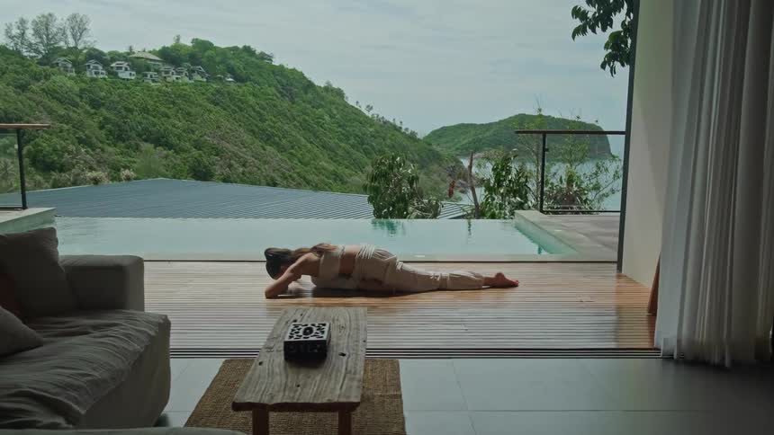 A tranquil woman gracefully performs a deep yoga stretch on the wooden deck of a tropical villa overlooking an infinity pool and the ocean