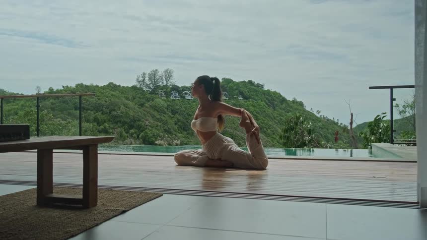 A serene woman holds a deep yoga stretch on a wooden deck by an infinity pool, with the camera slowly pulling back to reveal the luxurious villa and tropical hillside view