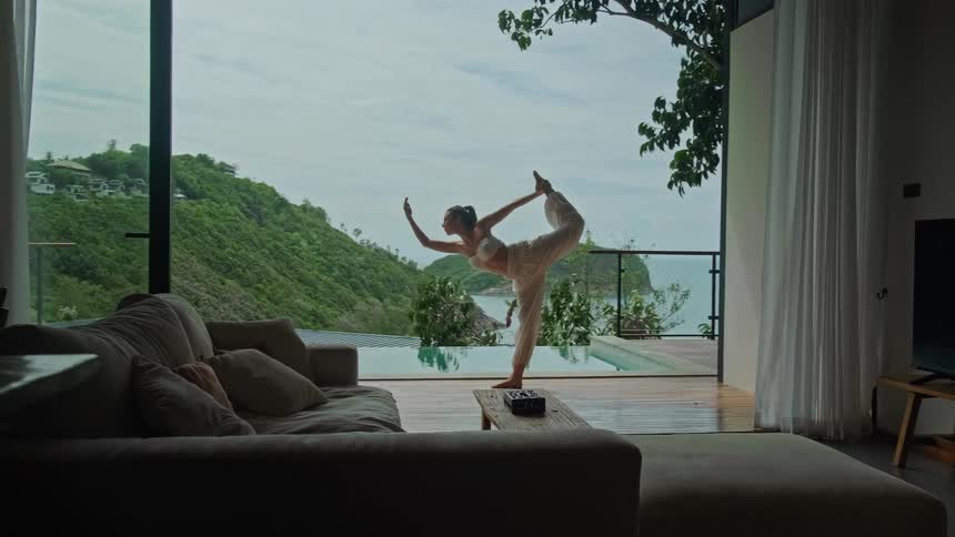 A woman gracefully practices a yoga pose on the deck of a luxurious villa overlooking an infinity pool and a beautiful tropical island seascape