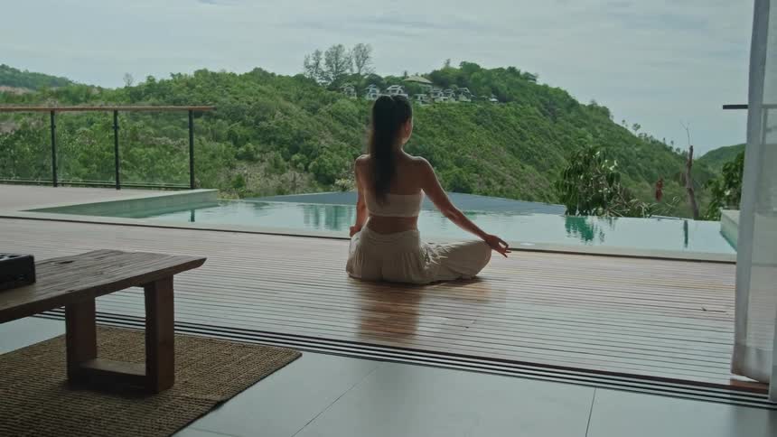Pulling back from a woman meditating in a lotus pose on a patio by an infinity pool to reveal a luxurious modern villa with a stunning mountain view