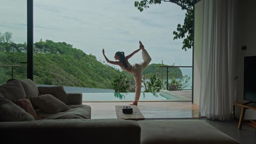 A woman practices a yoga balance pose on the deck of a luxury tropical villa with an infinity pool overlooking the ocean