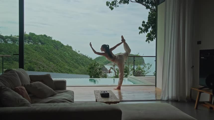 A woman gracefully holds a yoga pose on the deck of her luxury villa overlooking a stunning tropical ocean and hillside view
