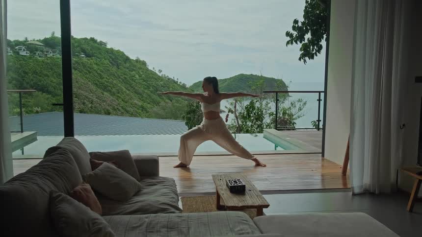 A woman holds a warrior yoga pose on a tranquil patio by an infinity pool with a stunning view of a tropical ocean bay