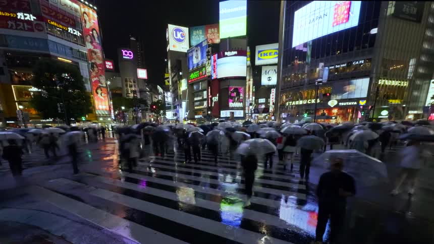 A rainy night time-lapse of the world-famous Shibuya Crossing in Tokyo, where crowds of people with umbrellas rush across the street under vibrant neon signs