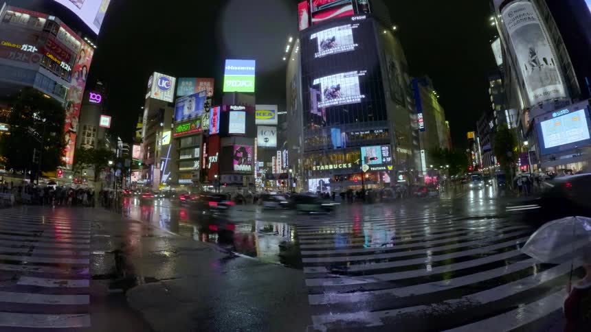 A wide-angle timelapse of the famous Shibuya Crossing in Tokyo on a rainy night, capturing the blur of crowds with umbrellas and traffic under vibrant neon lights