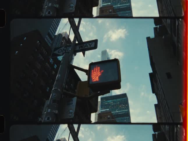 A vintage-style film captures a low-angle view of a red 'Don't Walk' street signal with New York City skyscrapers against the sky