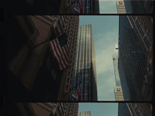 A nostalgic, upward-looking view of towering city skyscrapers reveals an American flag, captured on authentic vintage film