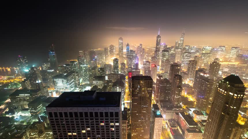 A dazzling night timelapse from a high vantage point shows the sprawling Chicago city skyline illuminated by a warm glow and the streaks of traffic below