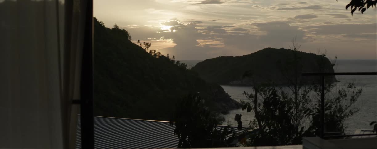 A tranquil view from a balcony shows a warm sunset over a tropical ocean bay surrounded by lush, silhouetted hills