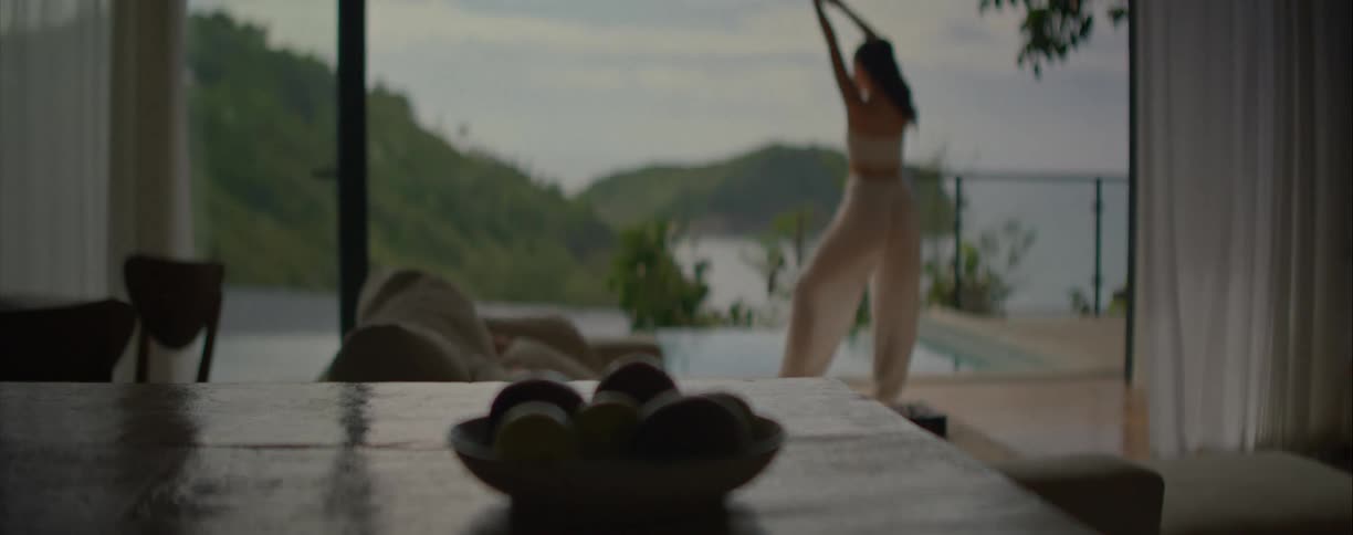 A slow focus pull from a fruit bowl reveals a woman dancing gracefully on a villa patio with a stunning tropical ocean view