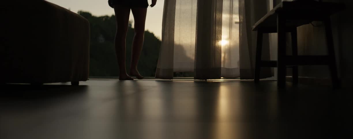 A silhouetted woman walks barefoot towards an open window where sheer curtains blow gently in the breeze during a golden sunset