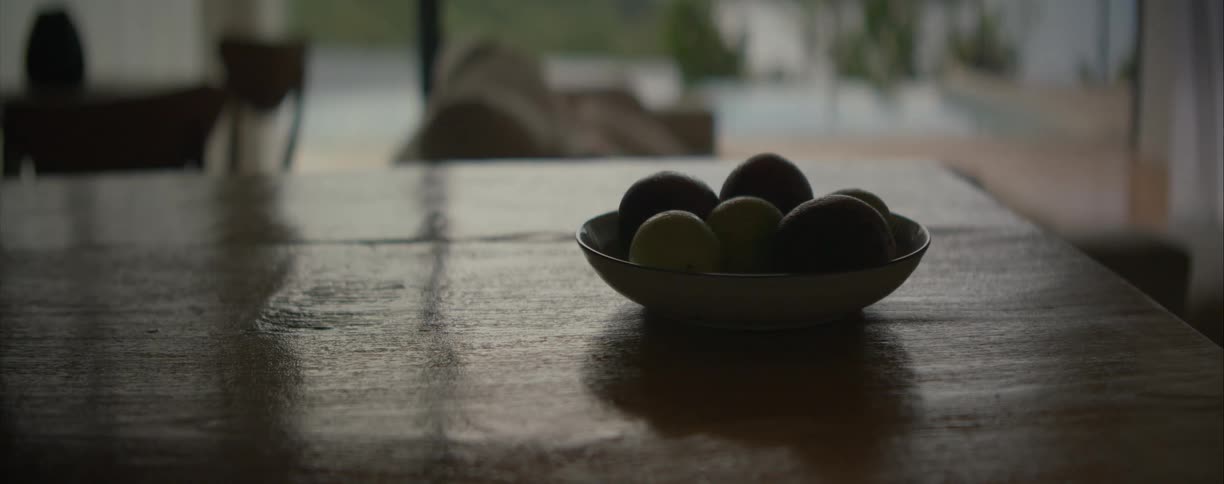 A moody still life shot of a fruit bowl resting on a reflective wooden table indoors