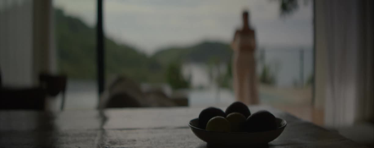 With a fruit bowl in the foreground, a woman gracefully dances on a tranquil balcony overlooking the sea at dusk
