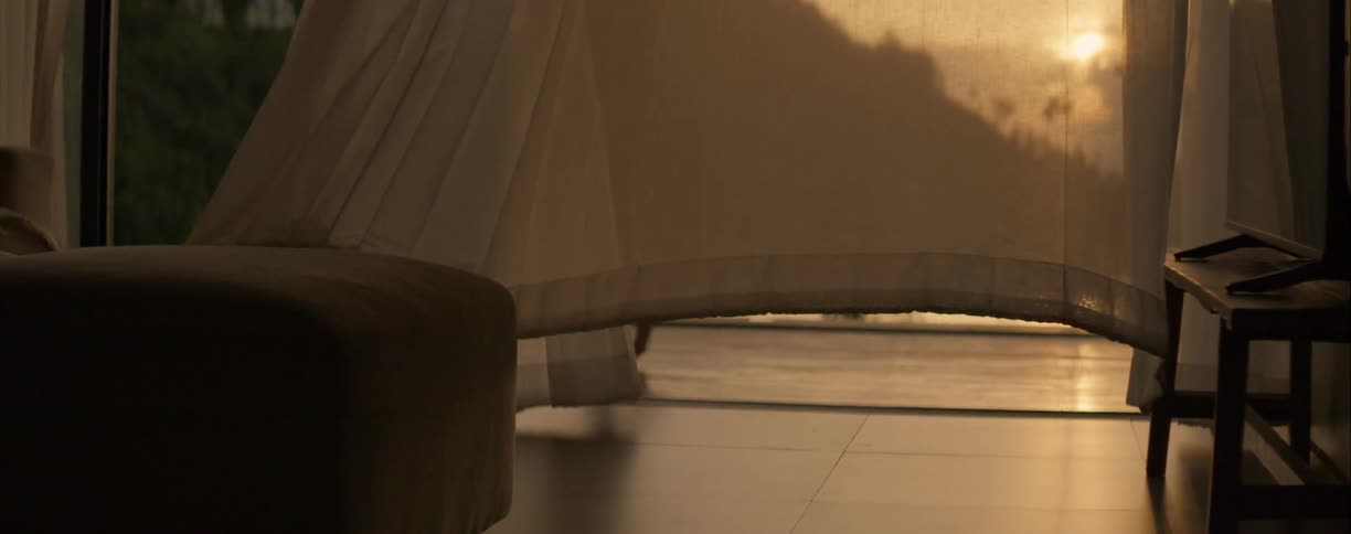 A woman's shadow is cast on a sheer curtain blowing in the breeze as she walks barefoot on a patio overlooking the water at sunset