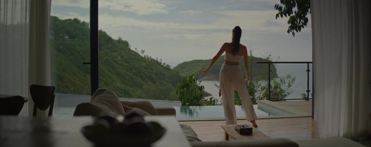 A woman in white gracefully performs a mindful dance routine by an infinity pool at a tropical villa overlooking the ocean