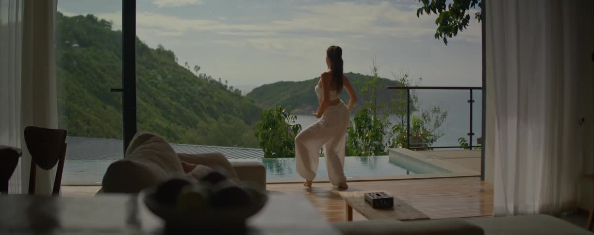 A barefoot woman in white performs a serene and expressive dance in slow motion on a beautiful villa's poolside deck, overlooking a lush tropical island