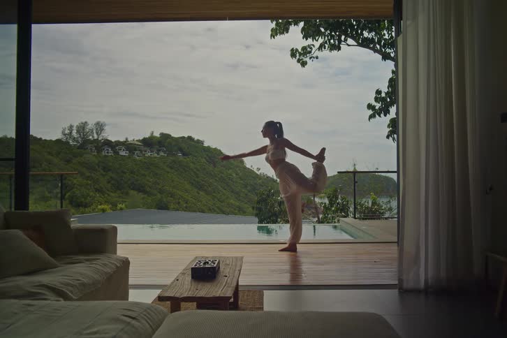 A woman gracefully practices a standing bow yoga pose on the deck of a modern villa, with an infinity pool overlooking a stunning tropical landscape