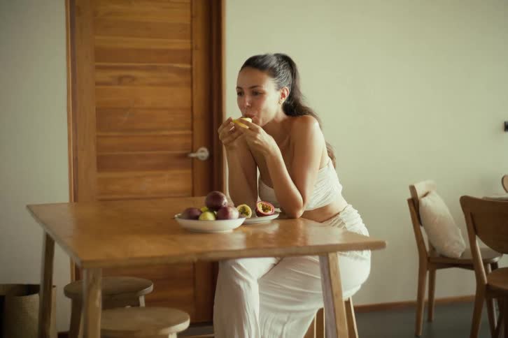A beautiful smiling woman dressed in white savors a fresh passion fruit for breakfast in a minimalist room
