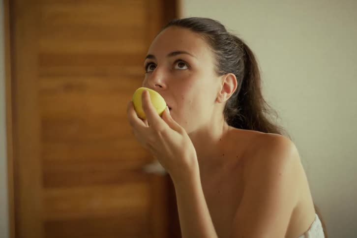 A beautiful young woman wrapped in a towel enjoys a healthy snack, eating a fresh passion fruit with a spoon in slow motion
