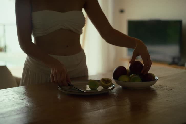 A woman in white carefully slices fresh passion fruit on a wooden table in a brightly lit room