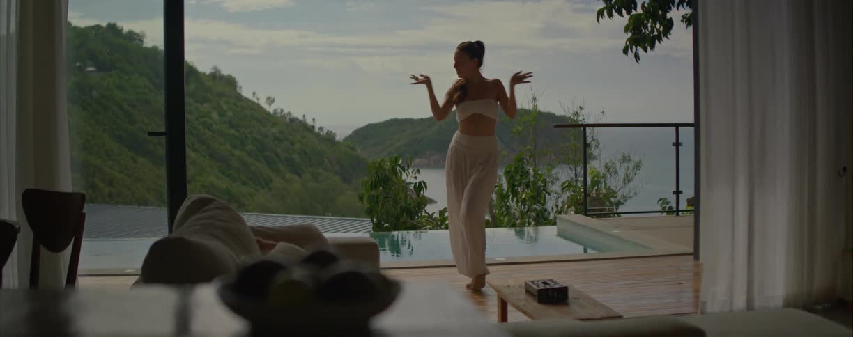 A woman in white clothing gracefully performs a slow, spiritual dance on a terrace with a stunning view of a tropical sea and hills
