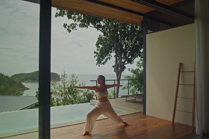 A serene woman performs yoga poses on a wooden deck of a luxurious tropical villa with a stunning ocean and infinity pool view
