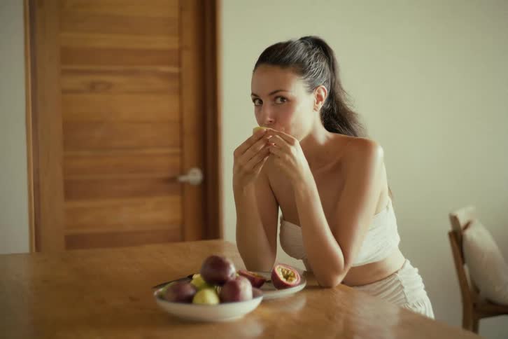 A beautiful woman in a white top sensually eats a fresh passion fruit at a wooden table in slow motion