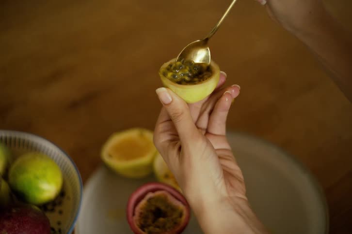 In an extreme slow-motion close-up, a woman's hand scoops the pulpy seeds from a fresh passion fruit with a golden spoon